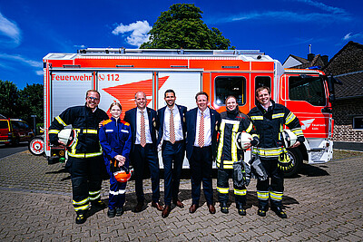 Das Dreigestirn steht in Anzug zwischen Feuerwehrmännern- und frauen vor einem Fahrzeug der Kölner Feuerwehr.