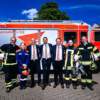 Das Dreigestirn steht in Anzug zwischen Feuerwehrmännern- und frauen vor einem Fahrzeug der Kölner Feuerwehr.