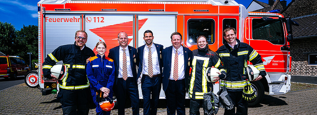 Das Dreigestirn steht in Anzug zwischen Feuerwehrmännern- und frauen vor einem Fahrzeug der Kölner Feuerwehr.