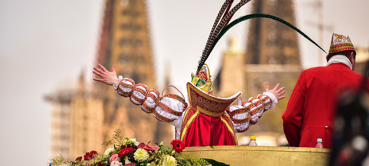 Der Prinz Karneval an Rosenmontag auf seinem Prunkwagen. Er streckt seine Arme jubelnd aus, der Dom ist im Hintergrund zu sehen.